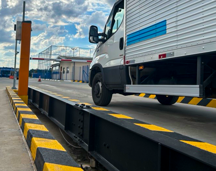 Caminhão leve sobre uma balança rodoviária em área industrial pavimentada. A estrutura da balança tem faixas amarelas e pretas e há uma guarita ao fundo, com céu parcialmente nublado.